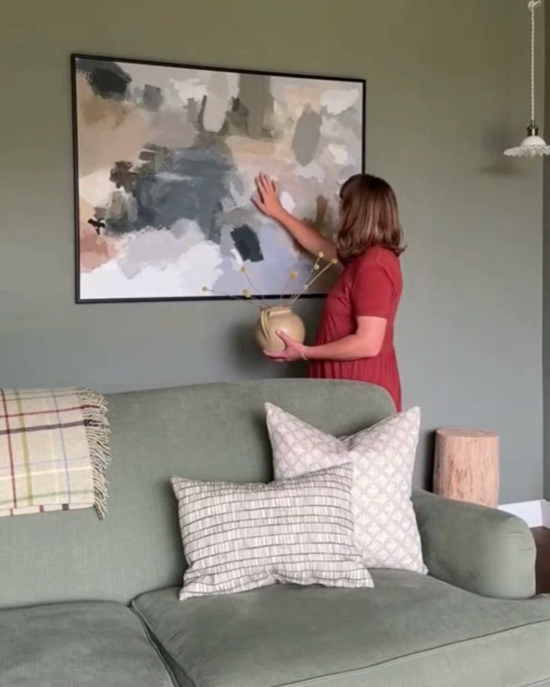 Woman hanging a large framed abstract painting above a green sofa