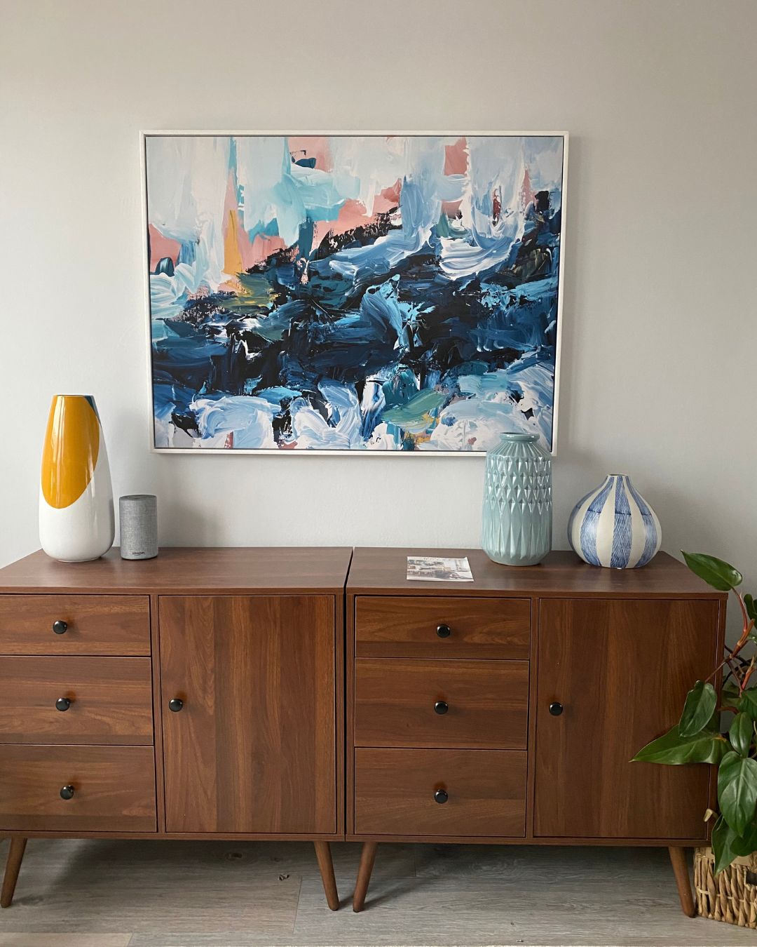 Blue abstract painting in a white frame above a walnut sideboard with vases